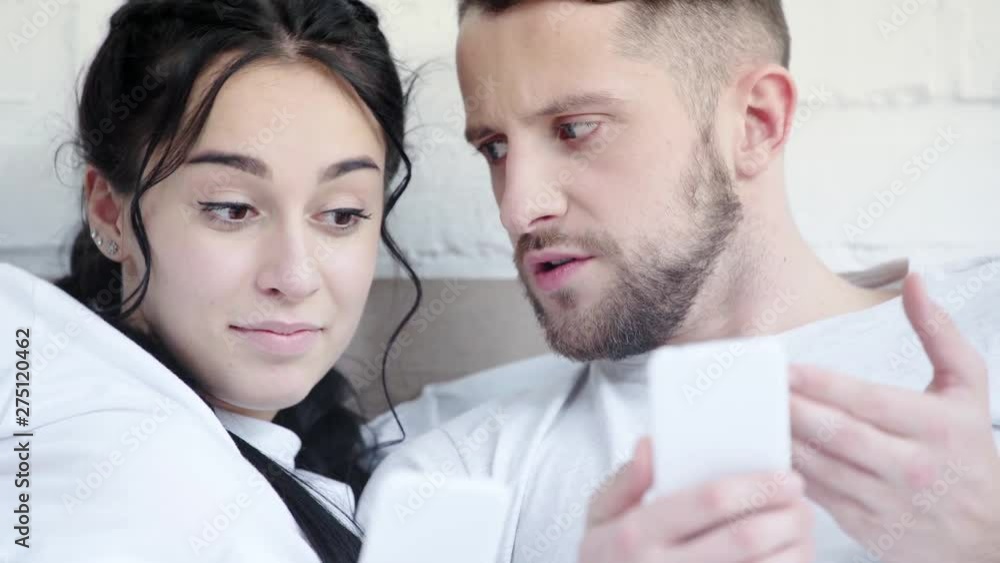 happy woman near dissatisfied boyfriend using smartphone and gesturing at home 