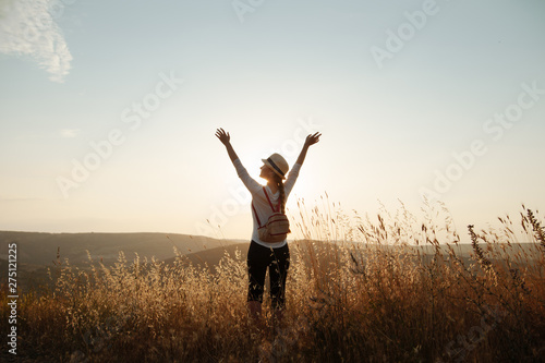 A young girl in a hat and a backpack stands on top of the mountain and raised her hands to the sky