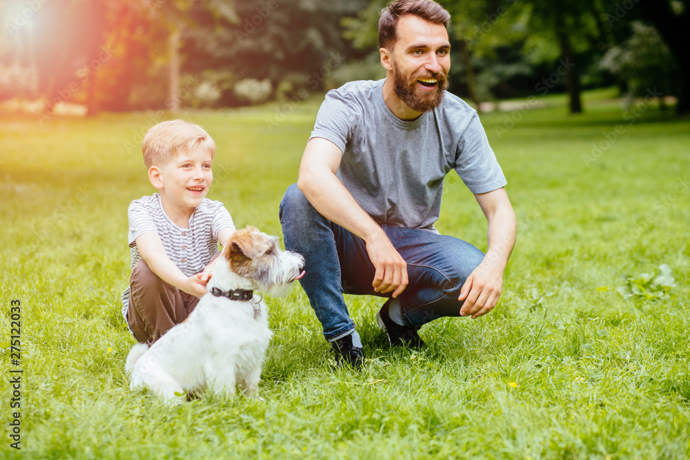 Fototapeta family, pet, domestic animal and people concept - happy family of father and schhol boy son happy laughing emotions on walk with dog Jack Russel Terrier in summer park. Sunglare effect.