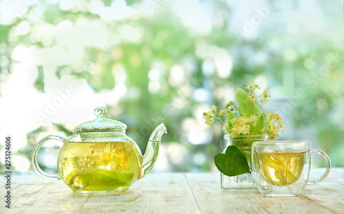 transparent glass teapot and cup with linden tea and flowers on white table in garden. Medicinal plant, flowers used for herbal teas, tinctures. herbal lime tree tea. close up. copy space