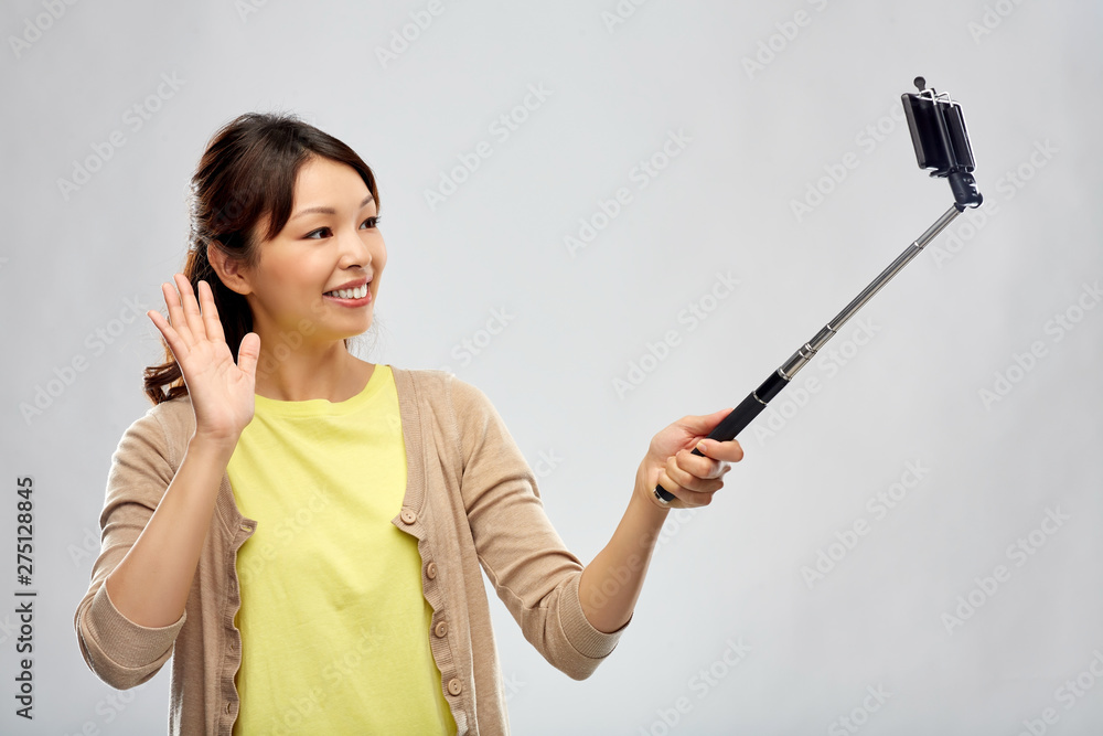 technology and people concept - happy asian woman taking picture by smartphone on selfie stick and waving hand over grey background technology and people concept - happy asian woman taking picture by smartphone on selfie stick and waving hand over grey background