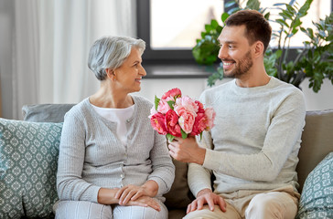 family, mother's day and birthday concept - smiling adult son giving flowers to his senior mother at home