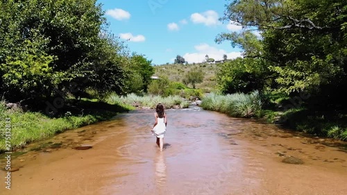 Wide shot of a woman in a white dress walking happily through a fresh water stream in the middle of the forest, normal speed version. Harmonious summer morning in the forest. photo