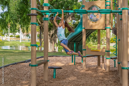 The young girl leaps and swings across the bars at the outdoor jungle gym. Focused on where to place her right hand as she hangs on to the top of the overhead bars at the playground.