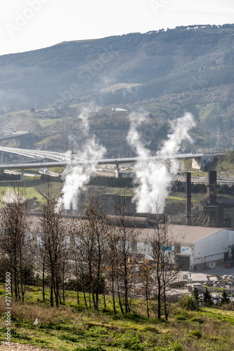 Industrial plant emitting smoke, gases, CO2, ... through chimneys in the middle of a green valley, on a sunny day