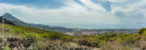 Stony path at upland surrounded by endemic plants. Sunny day. Clear blue sky and some clouds along the horizon line. Rocky tracking road in dry mountain area. Tenerife. Wide angle panorama