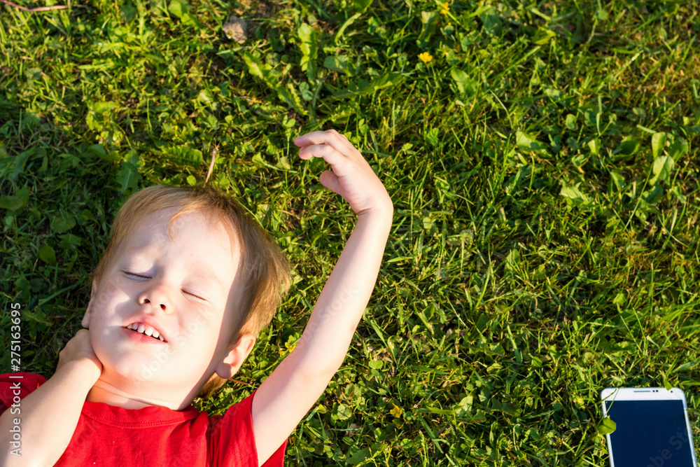 European child boy lies with his eyes closed on the grass and rests. The concept of education and dependence on gadgets in children. Relaxation with a smartphone European child boy lies with his eyes closed on the grass and rests. The concept of education and dependence on gadgets in children. Relaxation with a smartphone