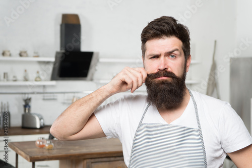 Curling his long moustache. Professional cook confident in his cooking skills. Bearded man during cooking in kitchen. Home-cooking and commercial cooking. The art of cooking