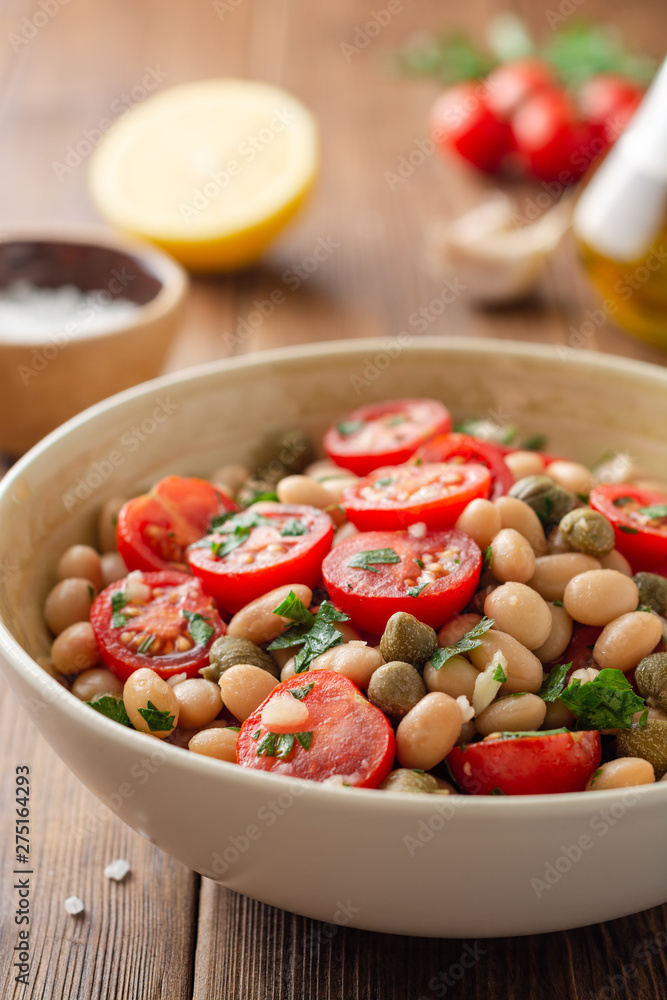 White bean salad with tomatoes, capers, garlic and parsley in bowl on wooden table. Selective focus. White bean salad with tomatoes, capers, garlic and parsley in bowl on wooden table. Selective focus.