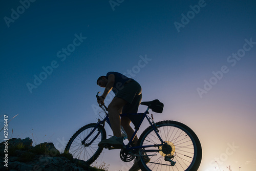 Silhouette of a fit male mountain biker riding his bike uphill on rocky harsh terrain on a sunset.