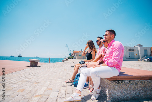 Four tourist friends having fun and taking pictures on a modern abstract bench paced on a seaside marine port on a sunny day. Enjoying the beautiful blue and turquoise sea. © qunica.com