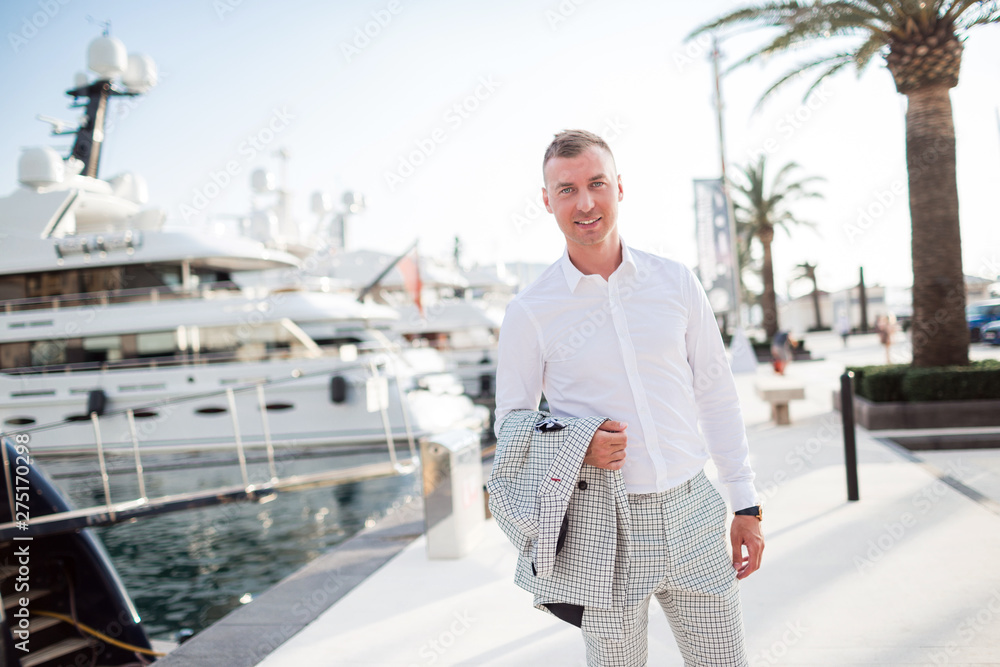 Φωτογραφία Stylish businessman is wearing white fashion costume stands on the background of the bay with luxury yachts