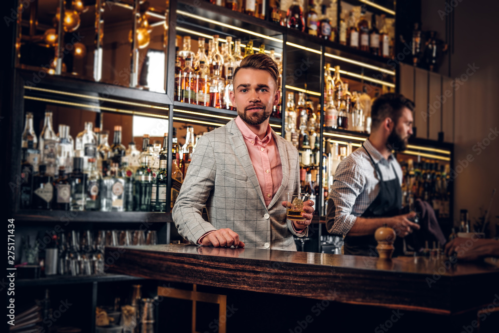 Handsome modern man in checkered suit is consuming alcohol at trendy bar.