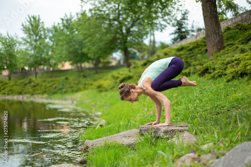 Slender Caucasian brunette girl doing yoga in summer on a green lawn by the river. woman doing Crane exercise, Bakasana pose