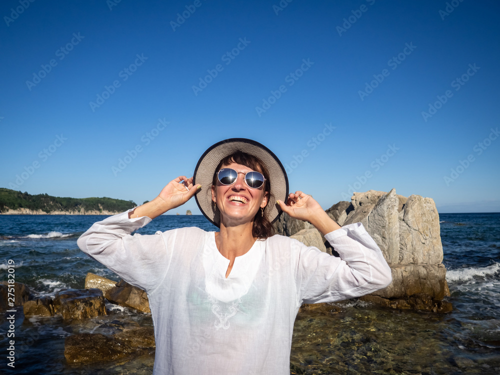 Vacation at sea: A girl in sunglasses and a straw slag on a rocky seashore in a bright sunny day. Bleak in the glasses.