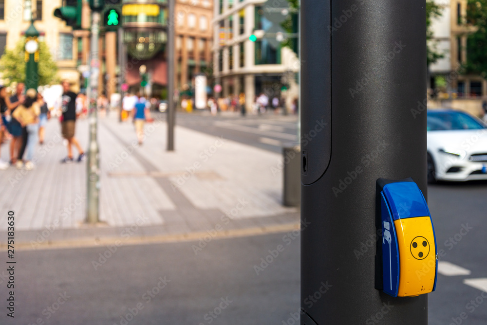 Yellow pedestrian crossing sign or crosswalk push button set up on metal post beside pedestrian crossing on the street in Germany with blur background of people stand, wait and walk to cross the road. Yellow pedestrian crossing sign or crosswalk push button set up on metal post beside pedestrian crossing on the street in Germany with blur background of people stand, wait and walk to cross the road.