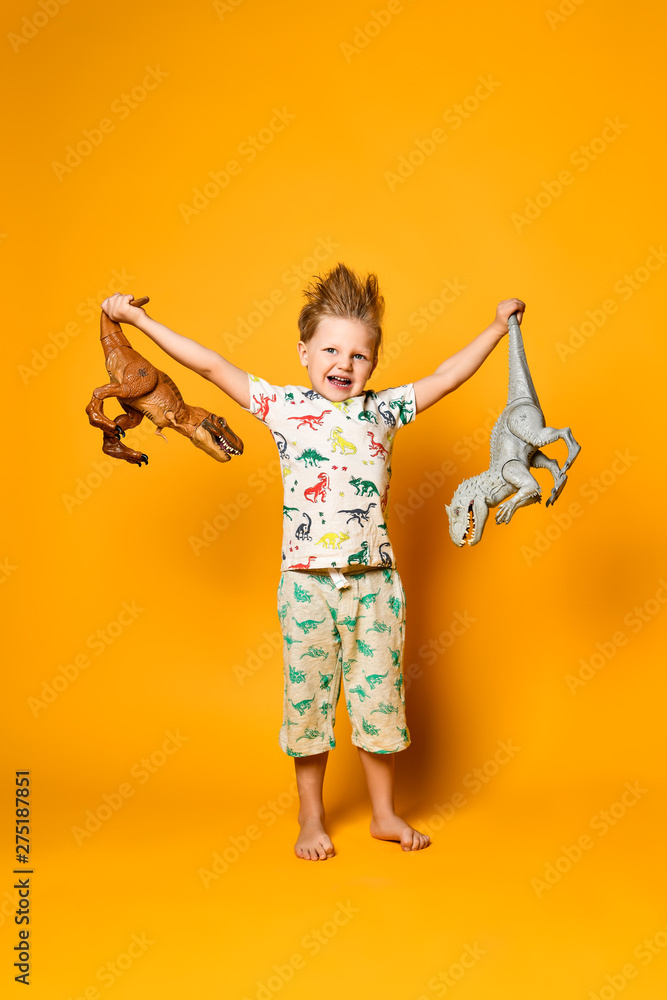 little cheerful boy holds a plastic dinosaur in each hand. The boy is dressed in a suit with pictures of dinosaurs. little cheerful boy holds a plastic dinosaur in each hand. The boy is dressed in a suit with pictures of dinosaurs.