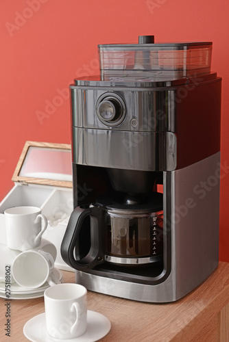 Modern coffee machine and cups on table against color background