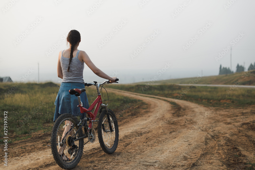white caucasian young woman in casual clothing walking with bike on village road, view from back in full body size, lifestyles stock photo image