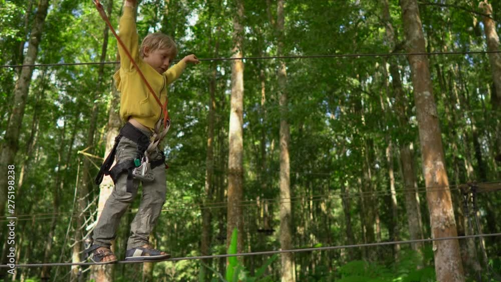 Little boy in a safety harness climbs on a route in a forest adventure park. He climbs on high rope trail. Outdoor amusement center with climbing activities consisting of zip lines and all sorts of Little boy in a safety harness climbs on a route in a forest adventure park. He climbs on high rope trail. Outdoor amusement center with climbing activities consisting of zip lines and all sorts of