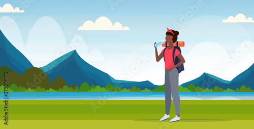 woman hiker with backpack drinking water african american girl traveler on hike hiking concept mountains landscape background full length flat horizontal