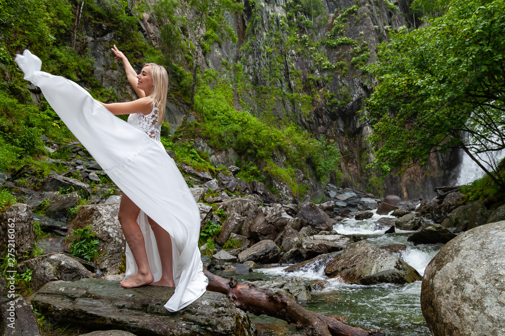 A young blonde girl in an elegant pose pulls up a boudoir dress in the mountains against a waterfall and stones raising her hands up like a ballerina on a warm summer day in Altai with green trees A young blonde girl in an elegant pose pulls up a boudoir dress in the mountains against a waterfall and stones raising her hands up like a ballerina on a warm summer day in Altai with green trees