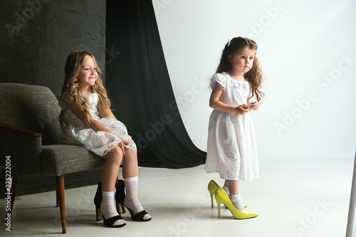 Interaction of children with adult world. Cute girls wearing mum's oversize shoes and dress for being older like she is. Little female models trying on clothes at home. Childhood, style, dream concept