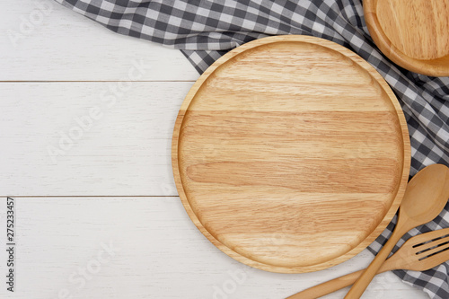 Empty round wooden plate with spoon, fork and grey gingham tablecloth on white wooden table. Top view image.