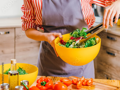 Healthy nutrition. Cropped shot of woman cooking, using kitchen tongs to mix vegetable salad in yellow bowl. photo