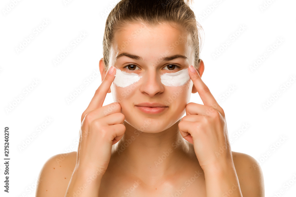 A young woman applying cream under her eyes on white background - obrazy, fototapety, plakaty 