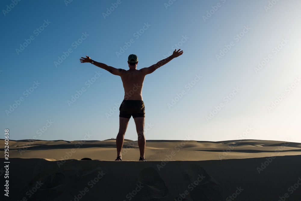 young man standing on top of sand desert dune with open arms as a symbol of being young and free and wanderlust