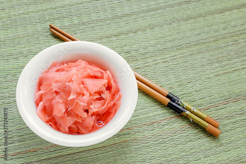 Pink pickled ginger in a white porcelain bowl and wooden chopsticks on a green wicker table mat. Traditional Japanese condiment for sushi. Healthy eating.