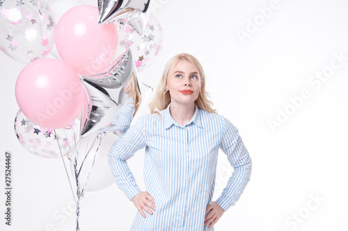 Portrait of dress of a cheerful beautiful girl is standing near a bunch of colorful balloons and celebrating isolated over a blue background