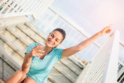 Beautiful happy young woman sitting on a steps and relaxing after a hard workout. Enjoying favorite song and using mobile phone. Exercising in urban environment
