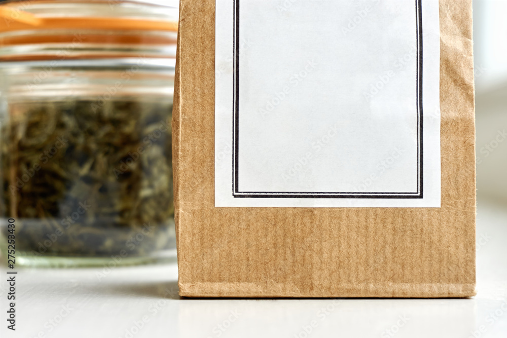 Fototapeta premium Brown kraft paper bag with white blank label in front of a bale glass jar and orange rubber gasket with dried herbs or tea on a white marble background. For use as a health template with copy space.