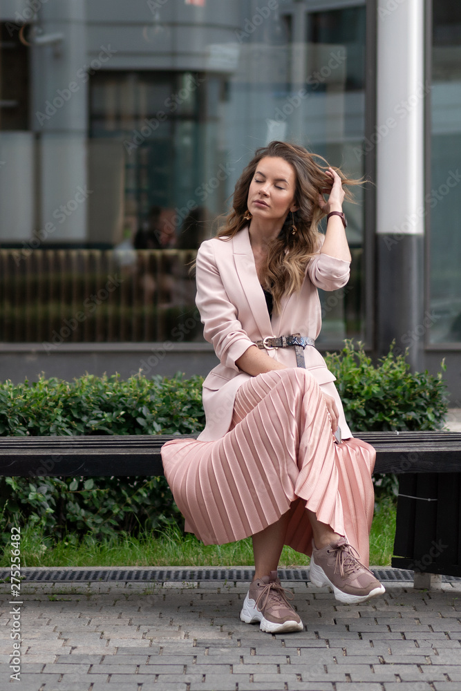 European girl sitting on a bench in a great mood. Cheerful posing with pleasure. Pink jacket, pleated skirt, sneakers. On the background of glass windows.