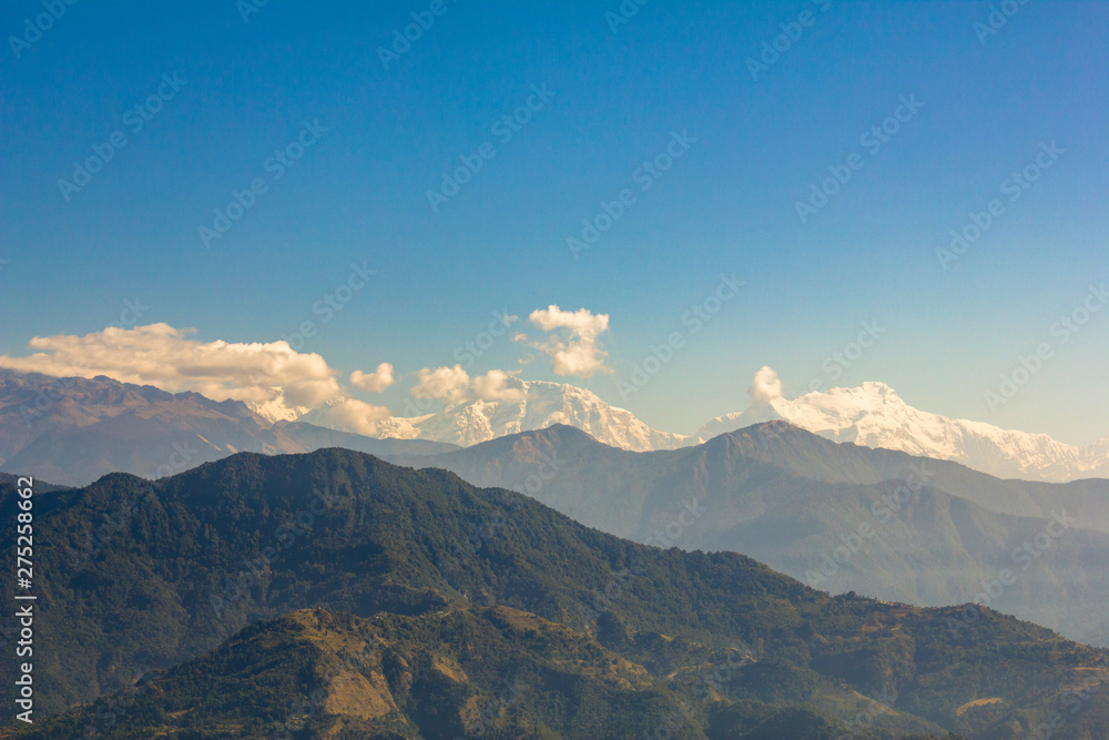 wooded green mountain in a haze against the snowy peaks of the Annapurna Ridge with white clouds under a clear blue sky