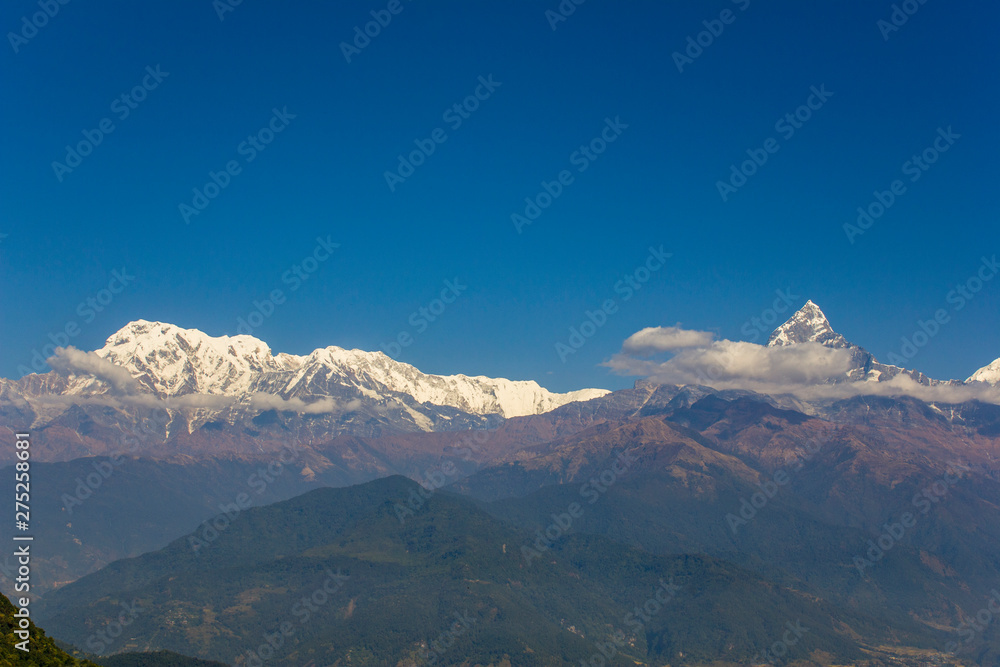 wooded hillsides in a mountain misty valley with snowy peaks of the Annapurna Ridge with white clouds under a clear blue sky