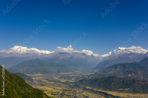 bright green slope of a hill against the backdrop of the city in a mountain valley and the snowy ridge of Annapurna under a clear dark blue sky