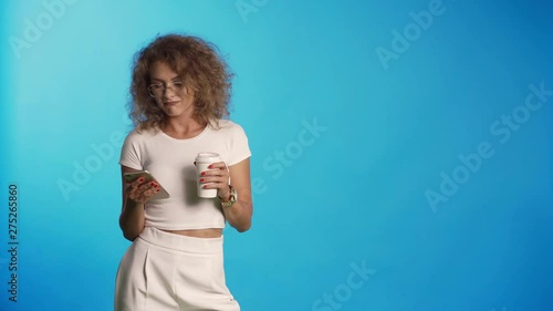 Happy woman use smartphone and drink coffee in studio on blue background. She are boored of news and trash in the feed photo