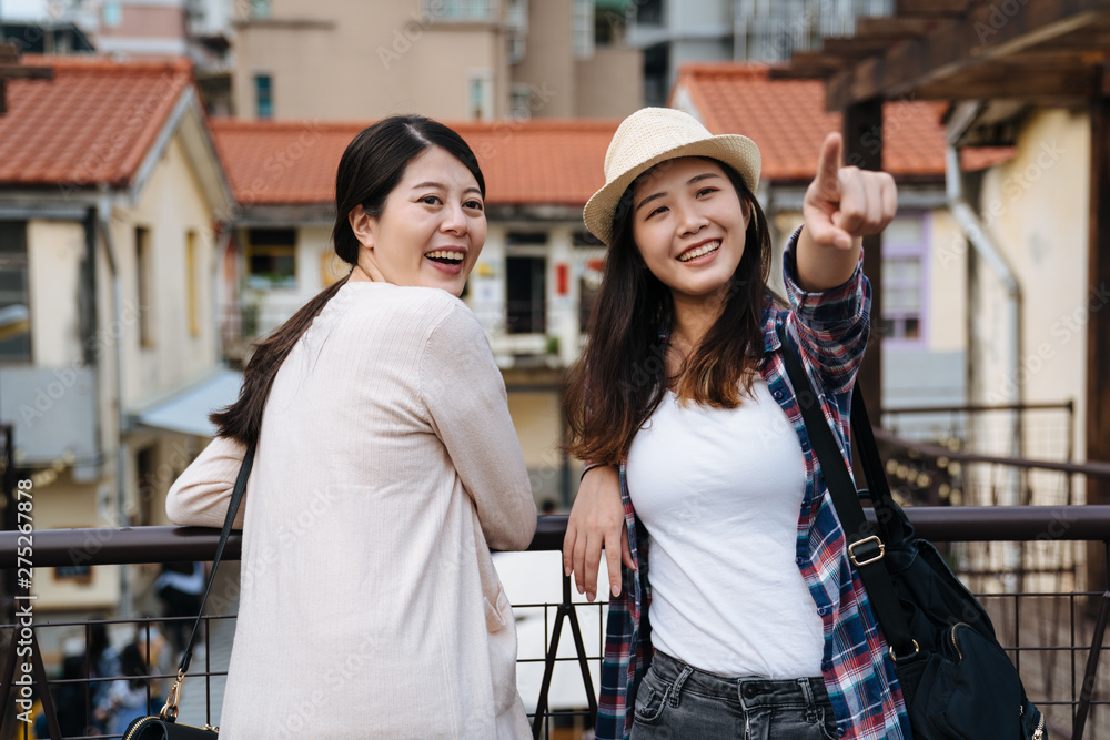 two young girls tourists travel in summer in japan small town. woman point at sky with beauitful sunset showing friend leaning on railing turn around in little village. smiling attractive ladies.