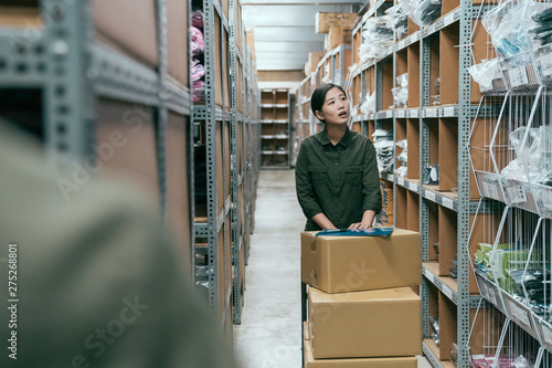 young asian female delivery postman pushing cart walking in storehouse of company. woman moving lots of cardboard boxes while doing stocktaking in warehouse. girl staff in uniform work in stockroom