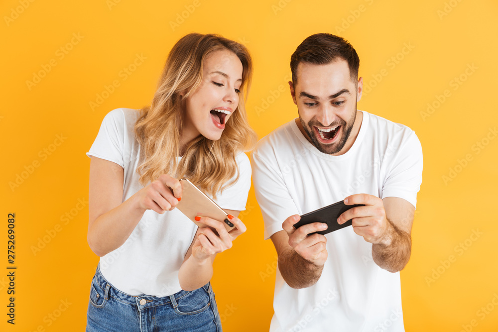 Portrait of happy young couple man and woman in basic t-shirts smiling and competing while playing video games on cellphones Portrait of happy young couple man and woman in basic t-shirts smiling and competing while playing video games on cellphones