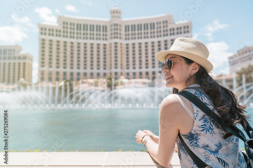 smiling young beautiful girl traveler standing near modern fountain with tall hotel building in background. woman tourist watching sights of seeing outdoor on sunny day in summer las vegas usa.
