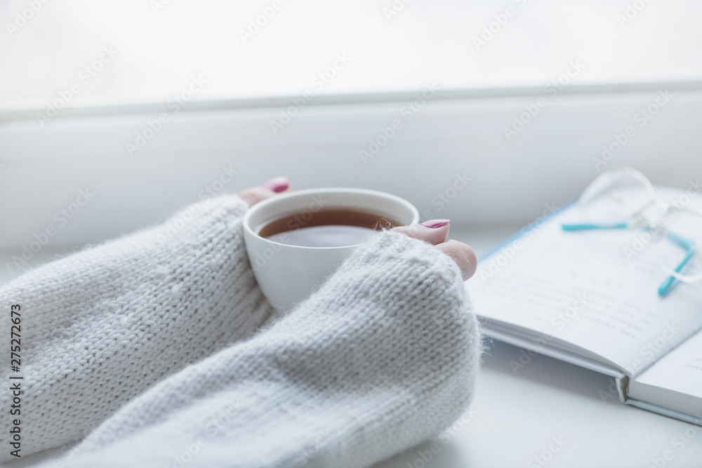 Tea drinking. The woman in a knitted, white pullover holds a cup of hot tea in her hands. Cozy weekends, winter drinks. Tea drinking. The woman in a knitted, white pullover holds a cup of hot tea in her hands. Cozy weekends, winter drinks.