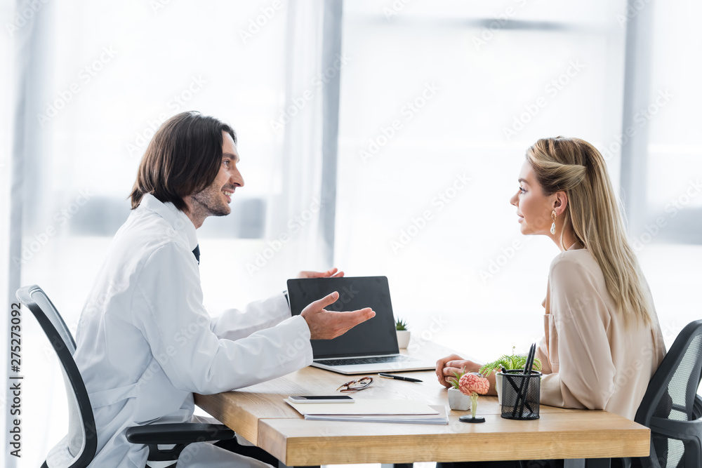 side view of cheerful doctor looking at patient and pointing with finger at laptop side view of cheerful doctor looking at patient and pointing with finger at laptop