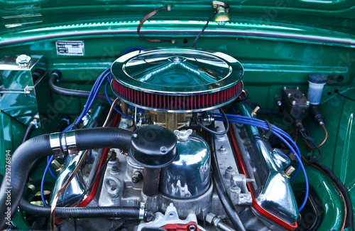 Classic muscle car engine close up view. Clean, shiny surface. Silver, chrome, red, blue and green colored part elements. Big round air intake with filter. Lots of tubes, wires, pipes, devices inside.