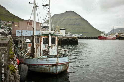 View on industrial harbour: Old, rusty, abandoned fishing boat moored in docks, big green hills, some of them covered with grey clouds in the background; Faroe Islands