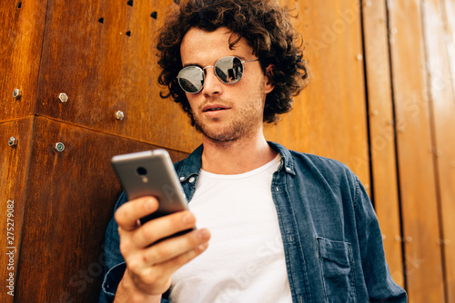 Close up portrait of handsome man standing outdoors, typing messages on mobile phone. Young male with curly hair wears sunglasses resting outside in the city browsing on his cell phone on modern wall
