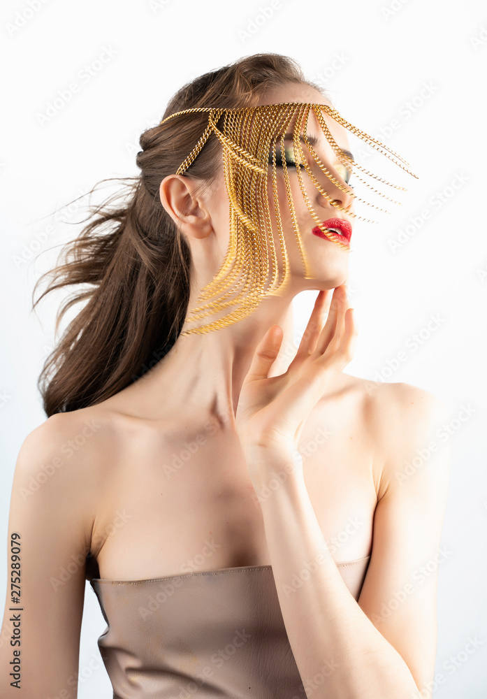 Naklejka premium Beautiful girl model with red lips make-up and naked shoulders wearing a conceptual fashion jewelry made of gold chains on her face, shakes her head, hair. Close up. Isolated on white.